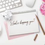 What’s stopping you – business concept. Motivational quote written on a ring binder, feminine styled modern workspace with coffee, crumpled paper balls, a bunch of flowers and office gadgets, top view
